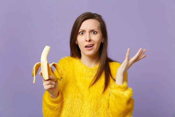 Perplexed puzzled young woman in fur sweater spreading hands, holding fresh ripe banana fruit isolated on violet pastel background. People vivid lifestyle, relax vacation concept. Mock up copy space.