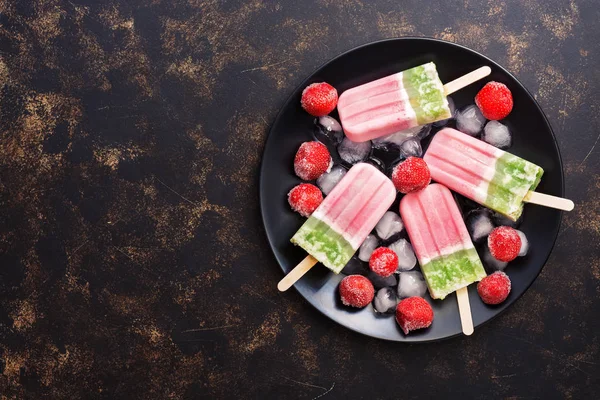 Strawberry ice cream on a stick with frozen berries on a black plate. Top view, copy space.