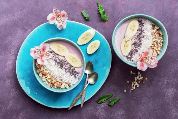 Fresh healthy breakfast - yogurt with banana, almond,coconut flakes, chia seeds in a blue bowl decorated with flowers on a purple background. View from above.