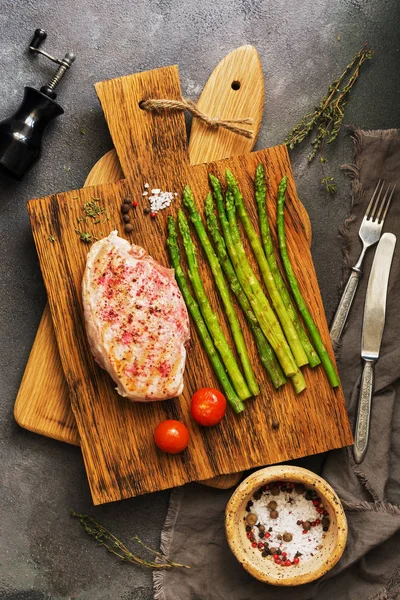 Grilled chicken fillet, asparagus, tomato and spices on a wooden cutting board, cutlery. Top view, flat lay.