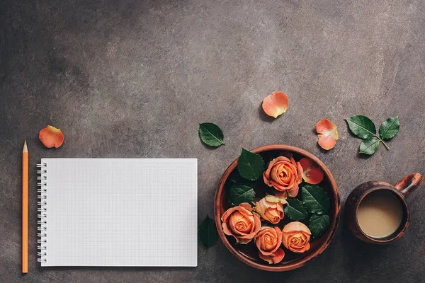 Female workspace with blank notepad, cup of coffee,ceramic bowl with flowers of coral roses and pencil. Top view, flat lay feminine beautiful background.