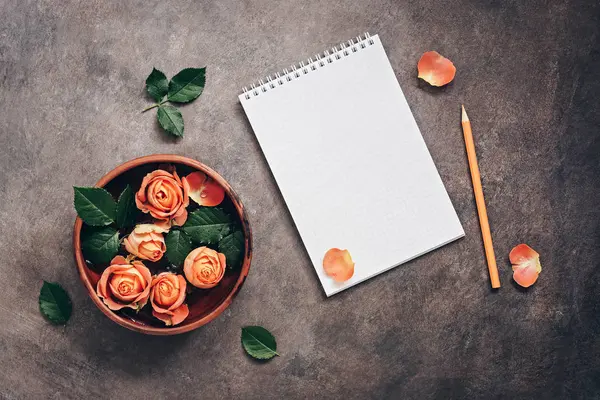 Blank notepad, ceramic bowl with flowers of coral roses and pencil on a dark rustic background.. Top view, flat lay feminine beautiful background.