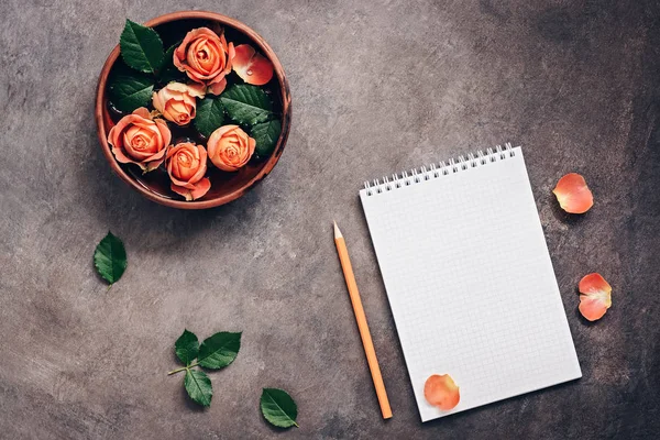 Blank notepad, flowers of coral roses and pencil on a dark rustic background.. Top view, flat lay feminine beautiful background.