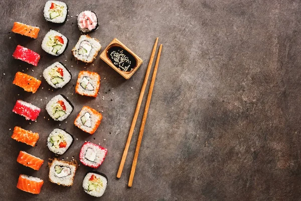 Flat lay rows of sushi rolls, soy sauce and chopsticks on a dark rustic background. View from above. Traditional asian food.