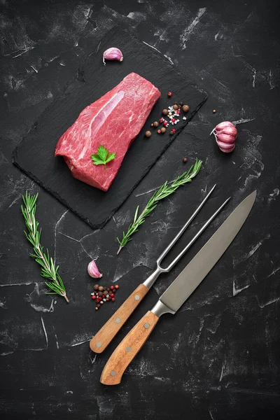 Raw beef steak with knife, fork and spices on a black stone background. Top view, flat lay