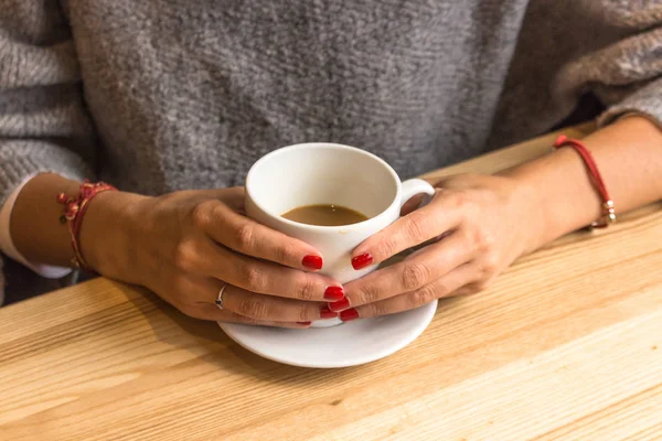 White cup of coffee in female hands. Warm your hands with a cup of hot drink. Meeting in a cafe. The girl in the coffee shop. Morning coffee in the restaurant. Red thread in the bracelet on hand. Red nail polish.