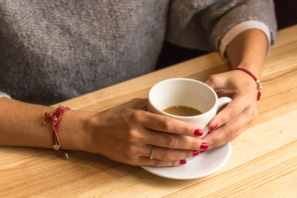 White cup of coffee in female hands. Warm your hands with a cup of hot drink. Meeting in a cafe. The girl in the coffee shop. Morning coffee in the restaurant. Red thread in the bracelet on hand. Red nail polish.