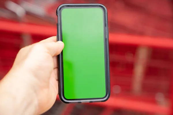 Man holding green screen smart phone with blurred shopping carts in background.