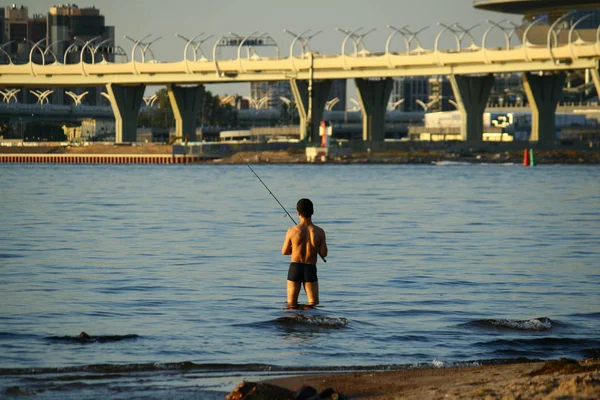 Um pescador com um torso nu pesca em uma vara de pesca no rio perto da ponte da cidade moderna — Fotografia de Stock