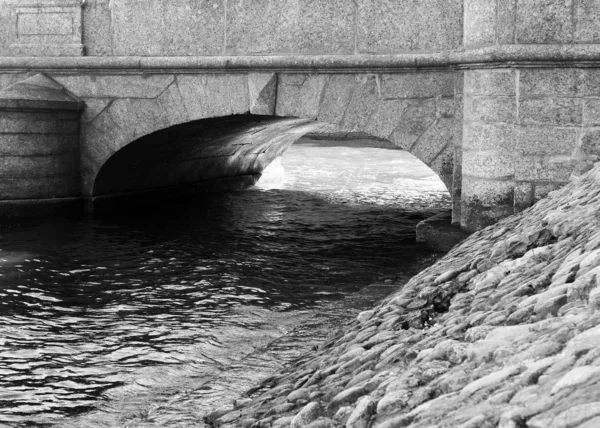Old stoned bridge over a small canal with stones on the beach in black and white. Bridge over a chanel of Neva river, Saint-Peterburg, Russia