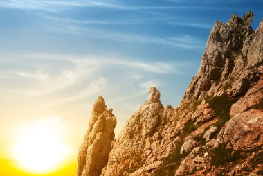 Akşam güneşinin ışığında kayalık dağ yamacı, gün batımı arka planı.