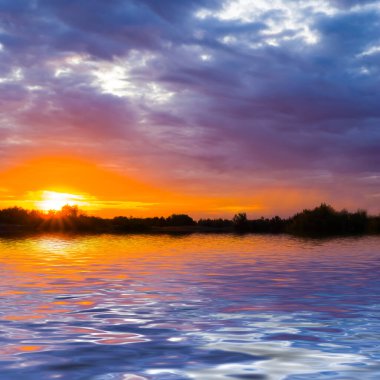 Sahilde orman siluetiyle gölün üzerinde dramatik bir gün batımı
