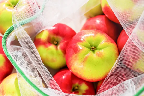 Fresh ripe and juicy apples in reusable eco-friendly mesh bag lie on the table. Sunny day. Plastic free, zero waste and sustainable lifestyle concept. Reusable using. Closeup view.