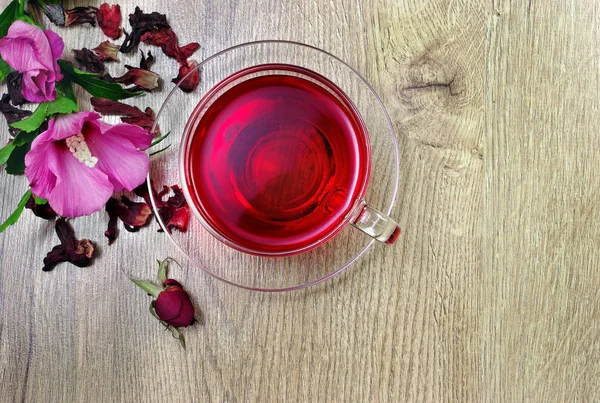 Hibiscus tea in a glass cup on a wooden table among the rose petals and dry tea. Hibiscus flower. Vitamin tea for cold and flu.