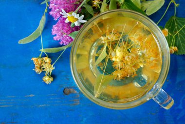 Eski mavi ahşap bir masada bir fincan ıhlamur çayı ve şifalı otlar. taze yonca, papatya ve ıhlamur. bitki çayı. grip ve soğuk algınlığı ilaçları.