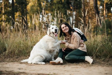Esmer kız orman yolunda beyaz golden retriever köpeğiyle oturuyor.