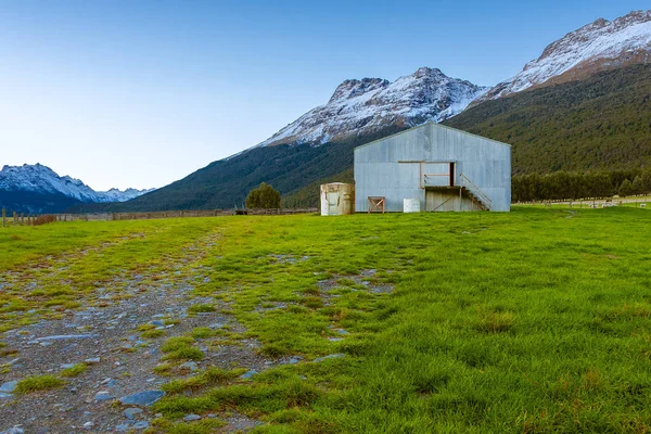 A barn on a grassland near mountains covered in snow, and a flock of sheep grazing nearby.