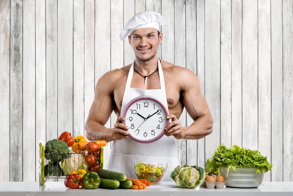 Man bodybuilder cooking on kitchen in white toque blanche and cook protective apron with clock