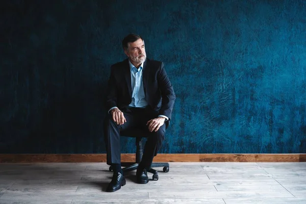 Retrato horizontal de atractivo hombre maduro serio con ropa formal elegante sentado en sillón y mirando a la cámara aislada sobre fondo azul oscuro . —  Fotos de Stock