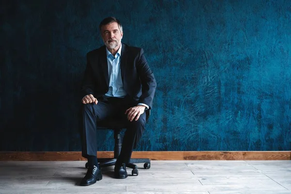 Retrato horizontal de atractivo hombre maduro serio con ropa formal elegante sentado en sillón y mirando a la cámara aislada sobre fondo azul oscuro . —  Fotos de Stock