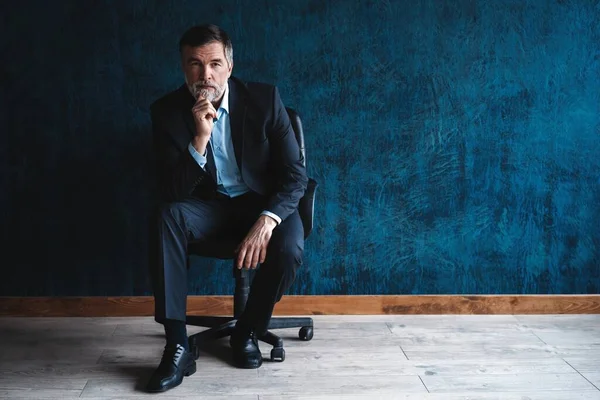 Retrato horizontal de atractivo hombre maduro serio con ropa formal elegante sentado en sillón y mirando a la cámara aislada sobre fondo azul oscuro . —  Fotos de Stock