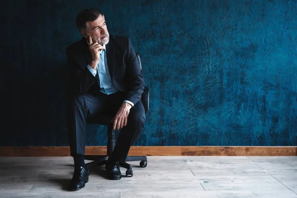 Horizontaal portret van aantrekkelijke serieuze volwassen man in stijlvolle formele kleding zittend in fauteuil en kijkend naar camera geïsoleerd over donkerblauwe achtergrond. — Stockfoto