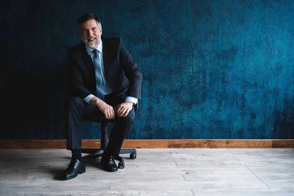 Retrato horizontal de atractivo hombre maduro serio con ropa formal elegante sentado en sillón y mirando a la cámara aislada sobre fondo azul oscuro . —  Fotos de Stock