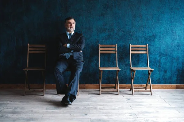 Ik wacht op een interview. Zelfverzekerde volwassen zakenman zittend op stoel tegen donkerblauwe achtergrond. — Stockfoto