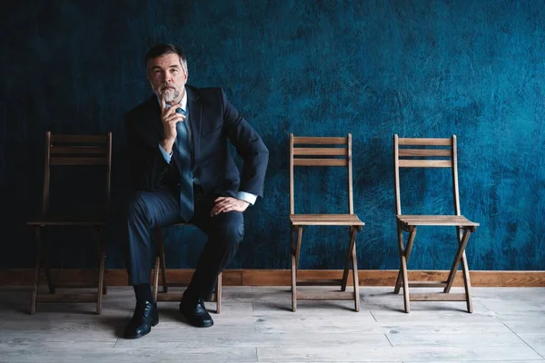 Waiting for interview. Confident mature businessman sitting on chair against dark blue background. — Stock Photo, Image