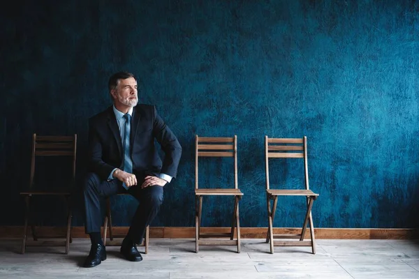 Ik wacht op een interview. Zelfverzekerde volwassen zakenman zittend op stoel tegen donkerblauwe achtergrond. — Stockfoto