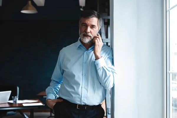 Volwassen zakenman praten op een mobiele telefoon staan door het raam met uitzicht op de stad. — Stockfoto