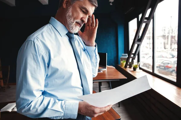 Gericht geconcentreerde accountant, volwassen zakenman kijkend naar het rapport, staat in vol ornaat, zo succesvol en intelligent, modern werkstation. — Stockfoto