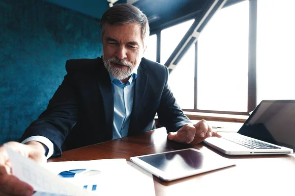 Ernstige volwassen financieel adviseur zittend aan de tafel en het controleren van financiële verslag met behulp van digitale tablet op kantoor. — Stockfoto