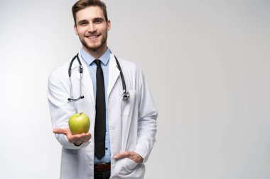 Beyaz önlüklü yakışıklı doktorun portresi kameraya bakıyor ve beyaz bir elmayı tutarken gülümsüyor..