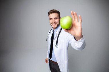 Beyaz önlüklü yakışıklı doktorun portresi kameraya bakıyor ve beyaz bir elmayı tutarken gülümsüyor..