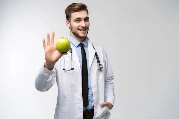 Portret van een knappe arts in witte jas die naar de camera kijkt en glimlacht terwijl hij een appel vasthoudt, geïsoleerd op wit. — Stockfoto