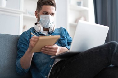 Evden çalışan genç beyaz bir adam koruyucu maske takıyor, dizüstü bilgisayar kullanıyor. Coronavirus salgını, covid 19 karantina