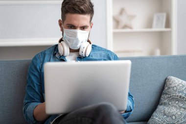 Evden çalışan genç beyaz bir adam koruyucu maske takıyor, dizüstü bilgisayar kullanıyor. Coronavirus salgını, covid 19 karantina