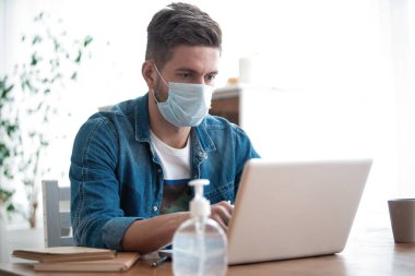 Evden çalışan genç beyaz bir adam koruyucu maske takıyor, dizüstü bilgisayar kullanıyor. Coronavirus salgını, covid 19 karantina
