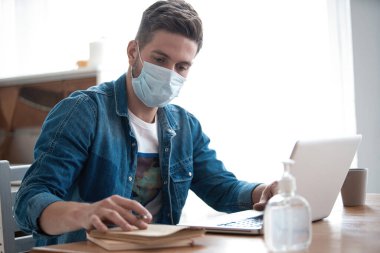 Evden çalışan genç beyaz bir adam koruyucu maske takıyor, dizüstü bilgisayar kullanıyor. Coronavirus salgını, covid 19 karantina