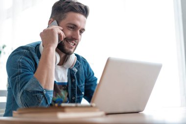 Evde oturma odasında çalışan sakallı adam. Laptop kullanan ve cep telefonuyla konuşan bir adam..
