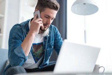 Evde oturma odasında çalışan sakallı adam. Laptop kullanan ve cep telefonuyla konuşan bir adam..
