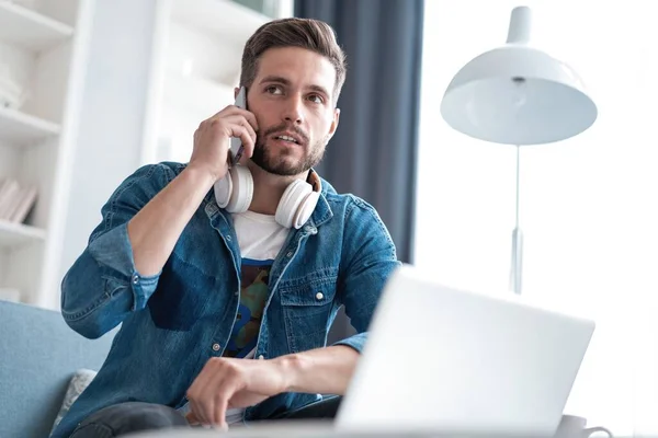 Hombre barbudo trabajando en la sala de estar en casa. Hombre usando el ordenador portátil y hablando en el teléfono móvil . — Foto de Stock