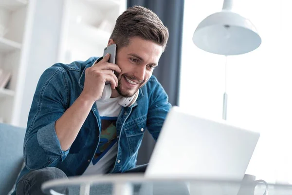 Hombre barbudo trabajando en la sala de estar en casa. Hombre usando el ordenador portátil y hablando en el teléfono móvil . — Foto de Stock