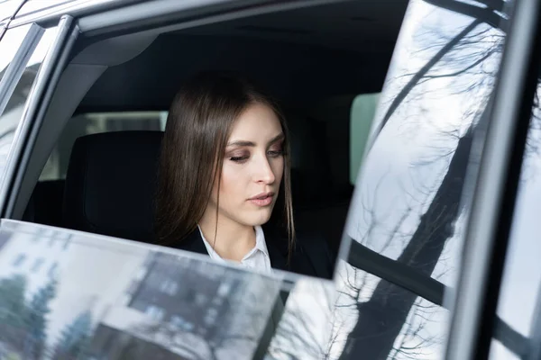 Zamyšlená mladá žena v obleku sedící na zadním sedadle auta a dívající se oknem. — Stock fotografie