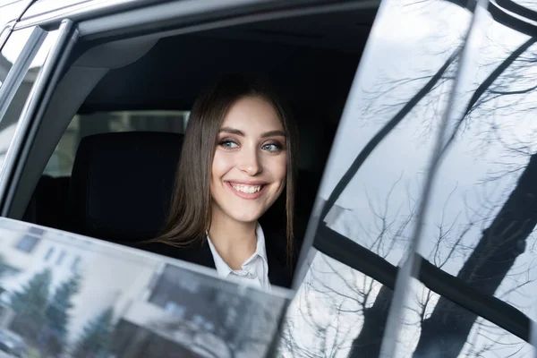 Mujer joven pensativa en un traje sentado en el asiento trasero de un coche y mirando a través de la ventana . —  Fotos de Stock