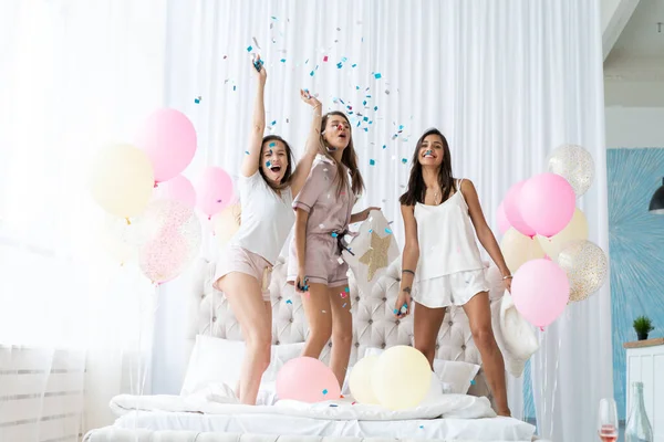Fyra attraktiva unga kvinnor i pyjamas ler och gestikulerar medan du hoppar in i sovrummet med konfetti flyger överallt — Stockfoto
