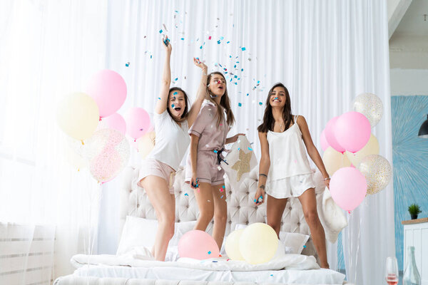 Four attractive young women in pajamas smiling and gesturing while jumping in bedroom with confetti flying everywhere