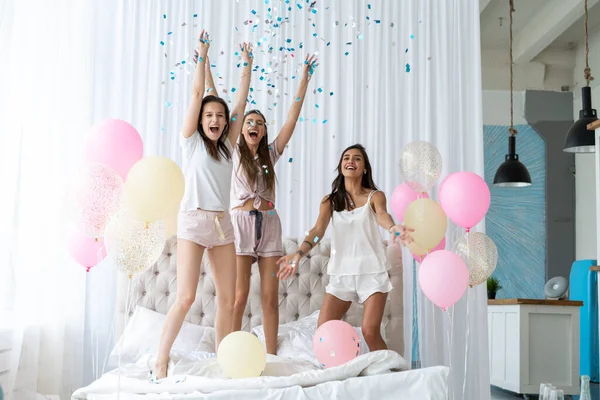 Четыре привлекательные девушки в пижаме улыбаются и жестикулируют, прыгая в спальню с конфетти, летающей повсюду — стоковое фото