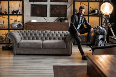 Deri ceketli yakışıklı genç adam taburede otururken gözlerini kaçırıyor.